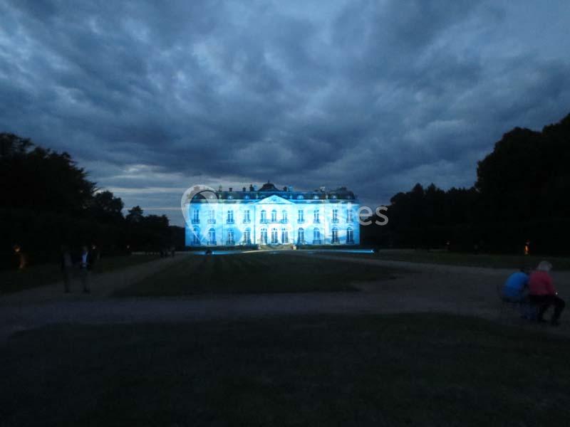 Façade d'un château illuminée en bleu sous un ciel nuageux, entourée de végétation et d'allées sombres.