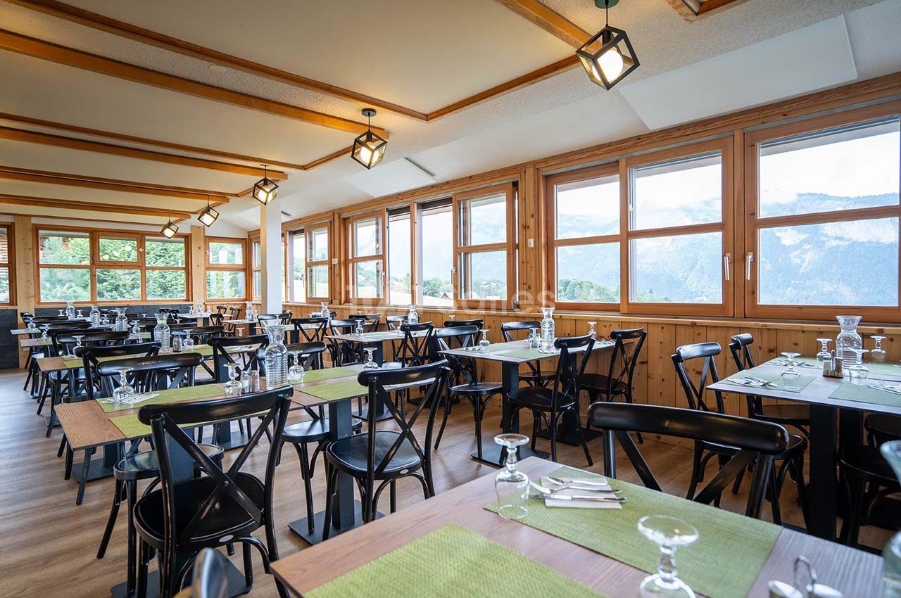 Salle de restaurant lumineuse avec tables dressées, grandes fenêtres offrant une vue sur les montagnes.