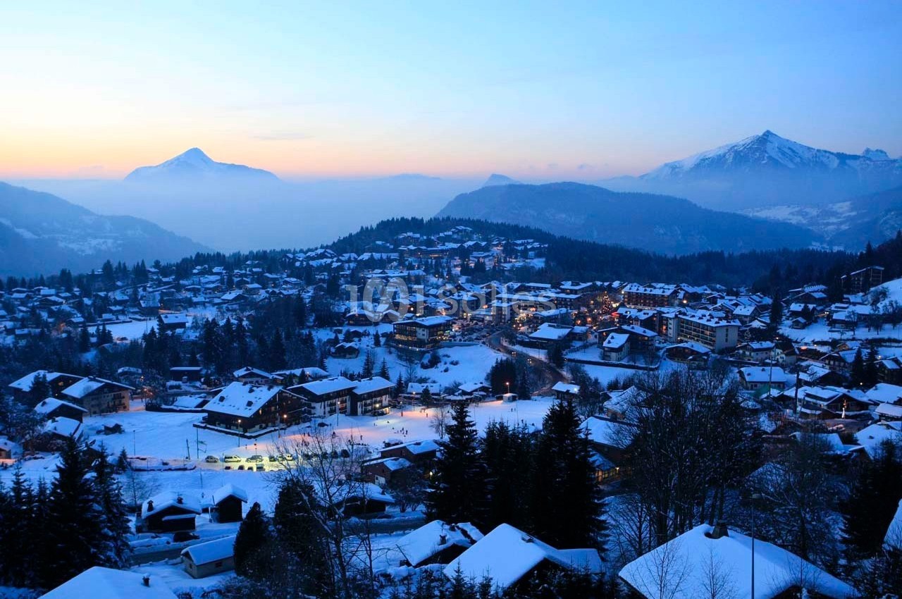 Vue d'un village de montagne enneigé au crépuscule, entouré de forêts et de sommets éclairés par une lumière douce.