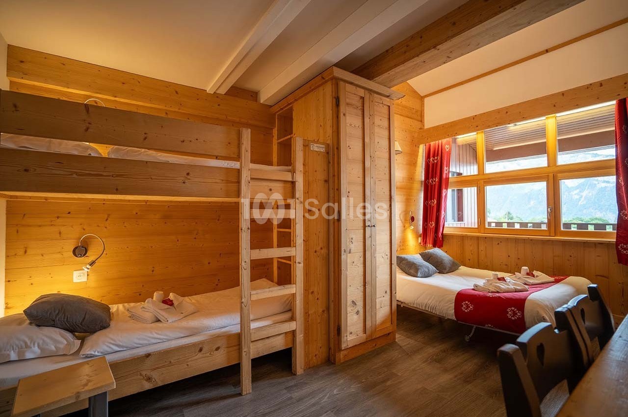 Chambre en bois avec lits superposés, lit double, fenêtre donnant sur une vue montagneuse et lumière naturelle.