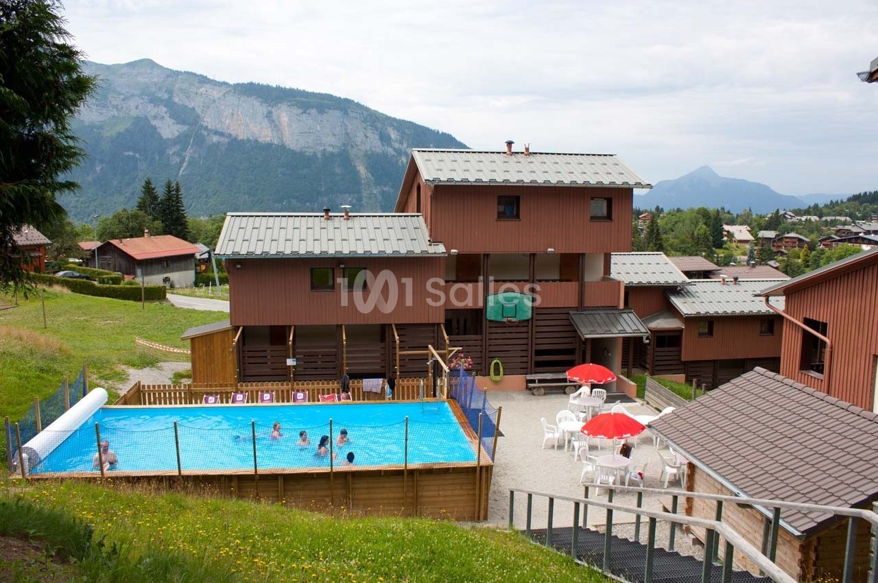 Piscine extérieure avec baigneurs, entourée de bâtiments en bois et de montagnes verdoyantes en arrière-plan.