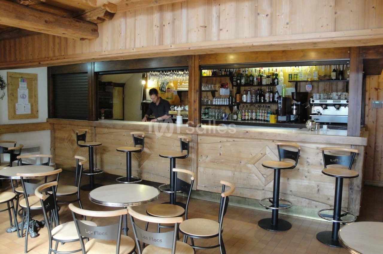Bar en bois avec tabourets hauts, tables rondes et étagères remplies de bouteilles, un serveur derrière le comptoir.