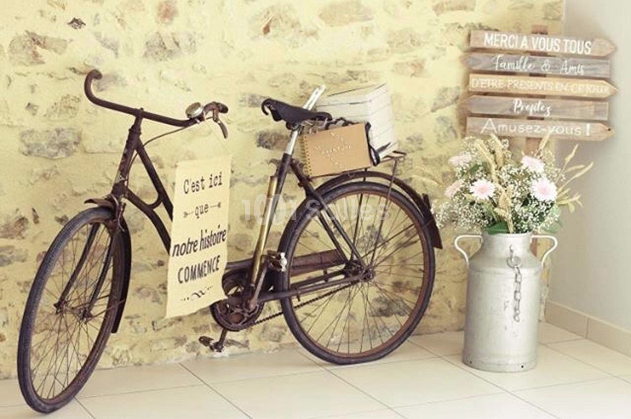 Un vieux vélo décoratif appuyé contre un mur en pierre, entouré de fleurs et d'une pancarte en bois.