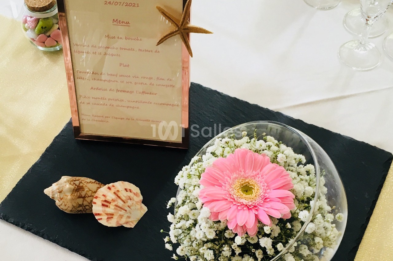 Menu encadré posé sur une table avec une décoration florale, des coquillages et des verres en arrière-plan.