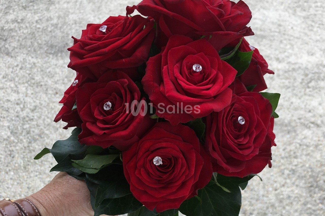 Bouquet de roses rouges ornées de perles brillantes, tenu par une main sur un fond gris clair.