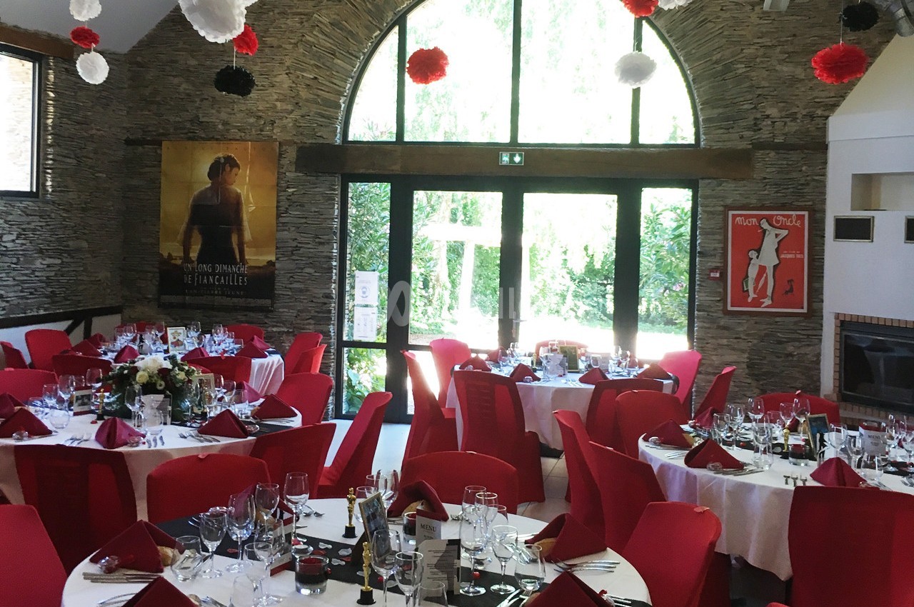 Salle de réception décorée avec des tables dressées, nappes blanches, serviettes rouges et affiches aux murs en pierre.