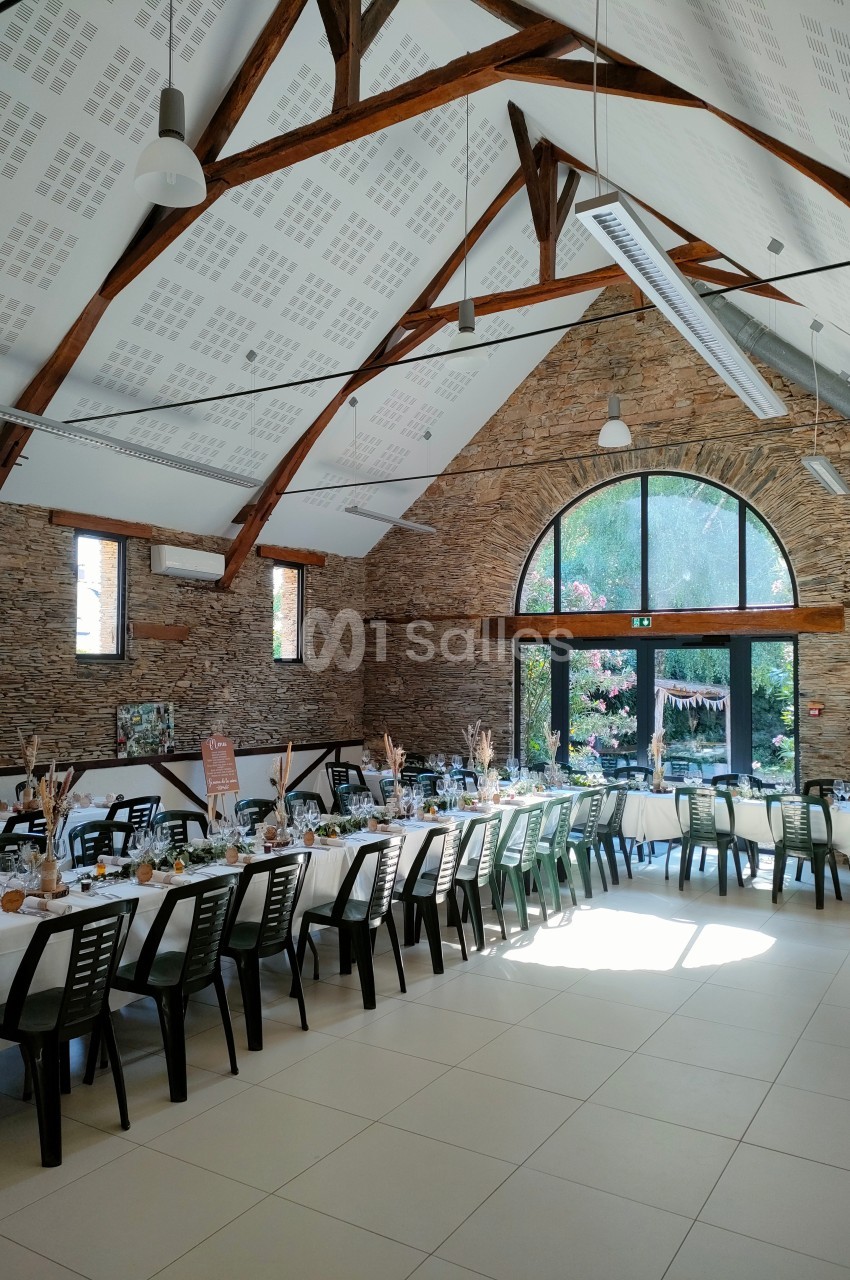 Salle de réception lumineuse avec tables décorées, poutres apparentes et grande baie vitrée donnant sur un jardin fleuri.