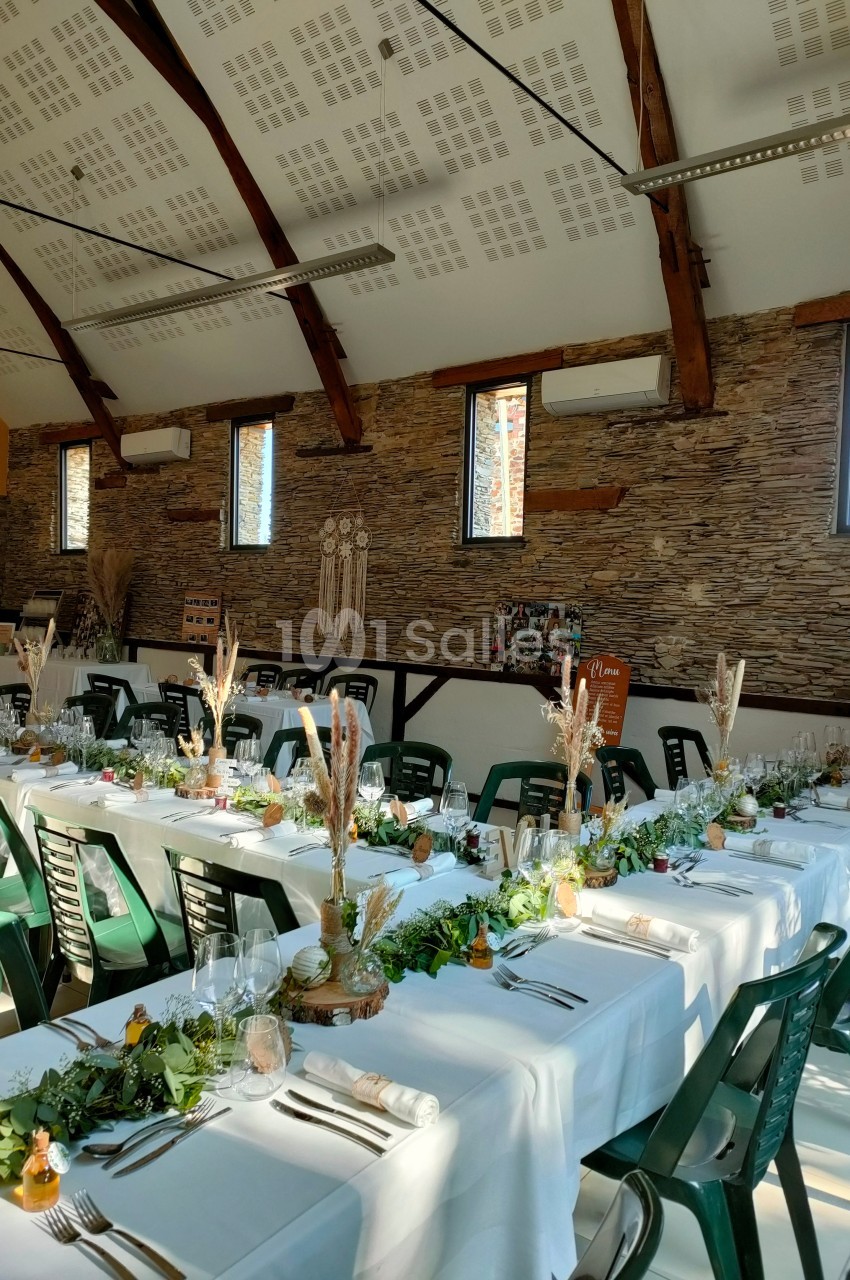 Salle de réception décorée avec des tables alignées, nappes blanches, centres de table végétaux et chaises vertes.