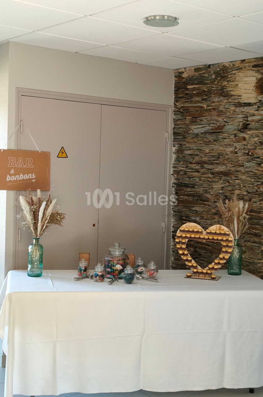 Table décorée avec un bar à bonbons, des pots en verre remplis de friandises et un cœur en bois sur un mur en pierre.