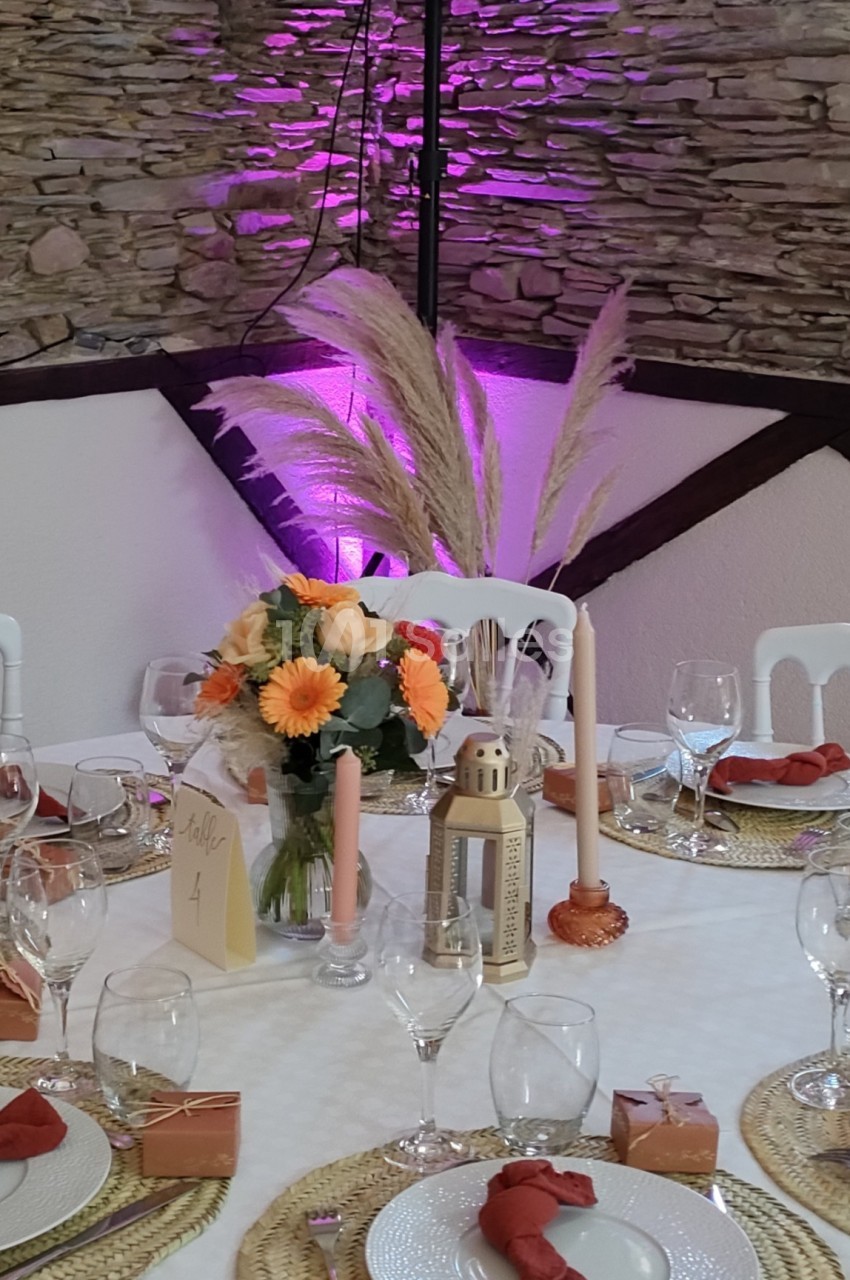 Table décorée pour un événement avec fleurs, bougies, lanternes et serviettes rouges, devant un mur en pierre éclairé.