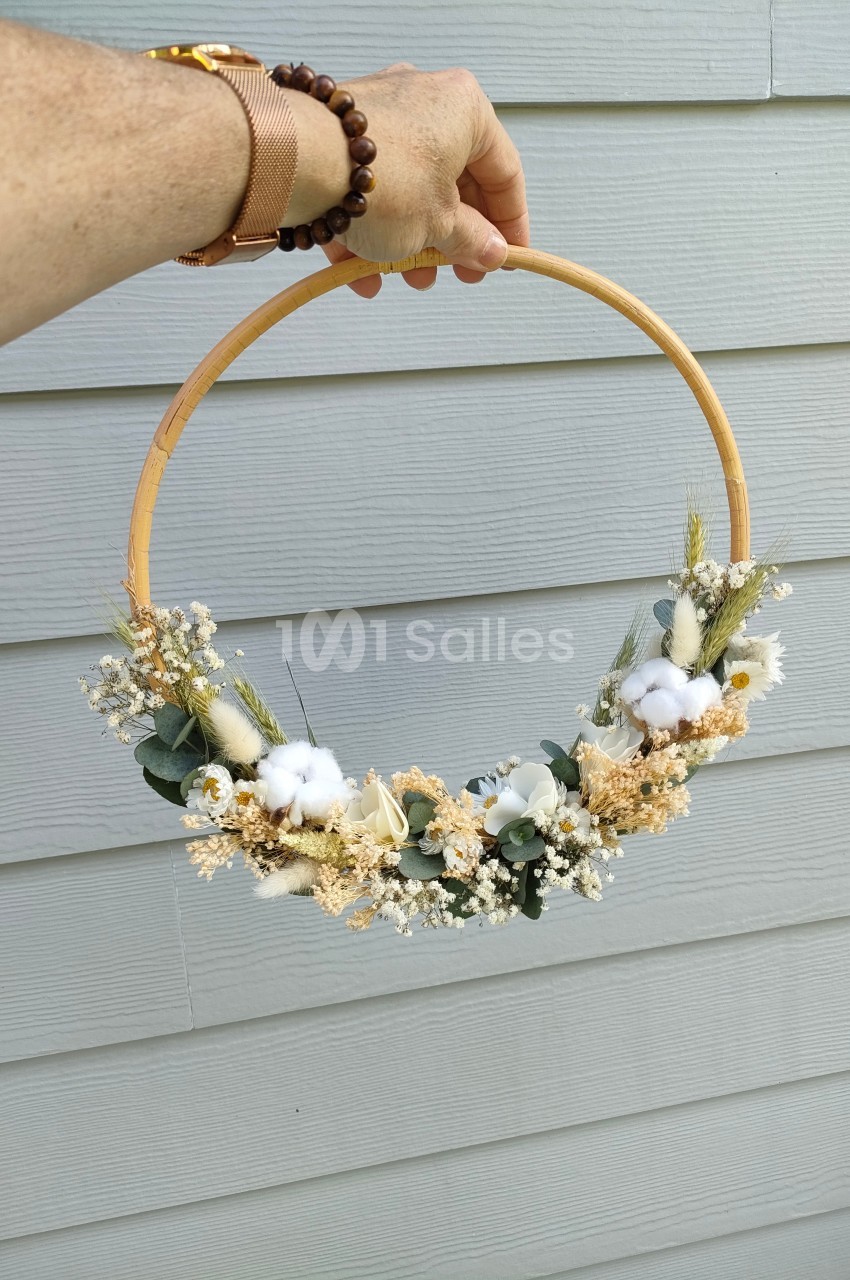 Couronne décorative en fleurs séchées et coton, tenue à la main devant un mur en bois gris.