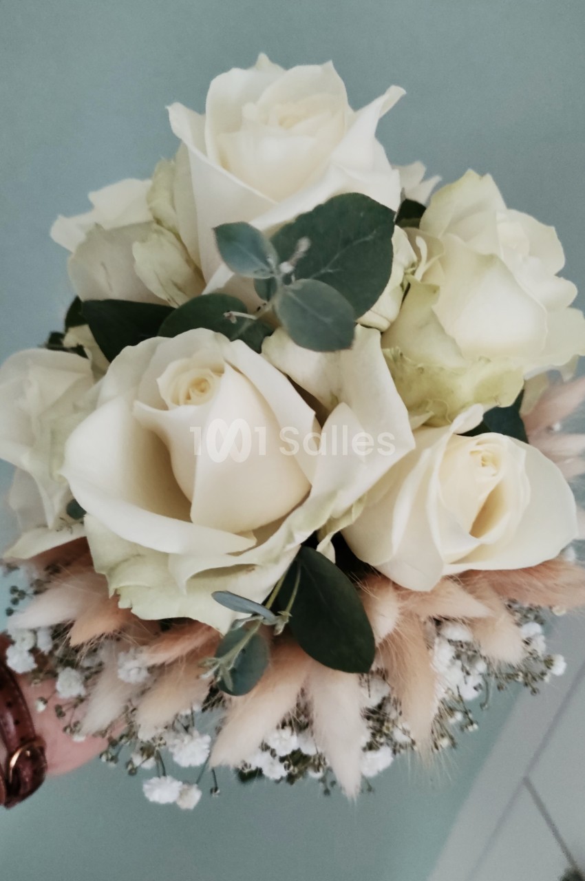 Bouquet de roses blanches entourées de feuillage vert, de gypsophile et de fleurs séchées beige, tenu à la main.