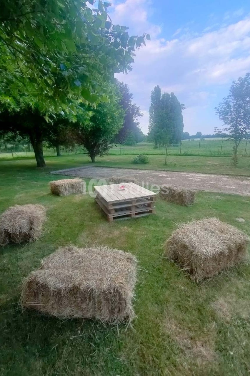 Bottes de paille disposées autour d'une table basse en palettes sur une pelouse, avec des arbres et un chemin en arrière…