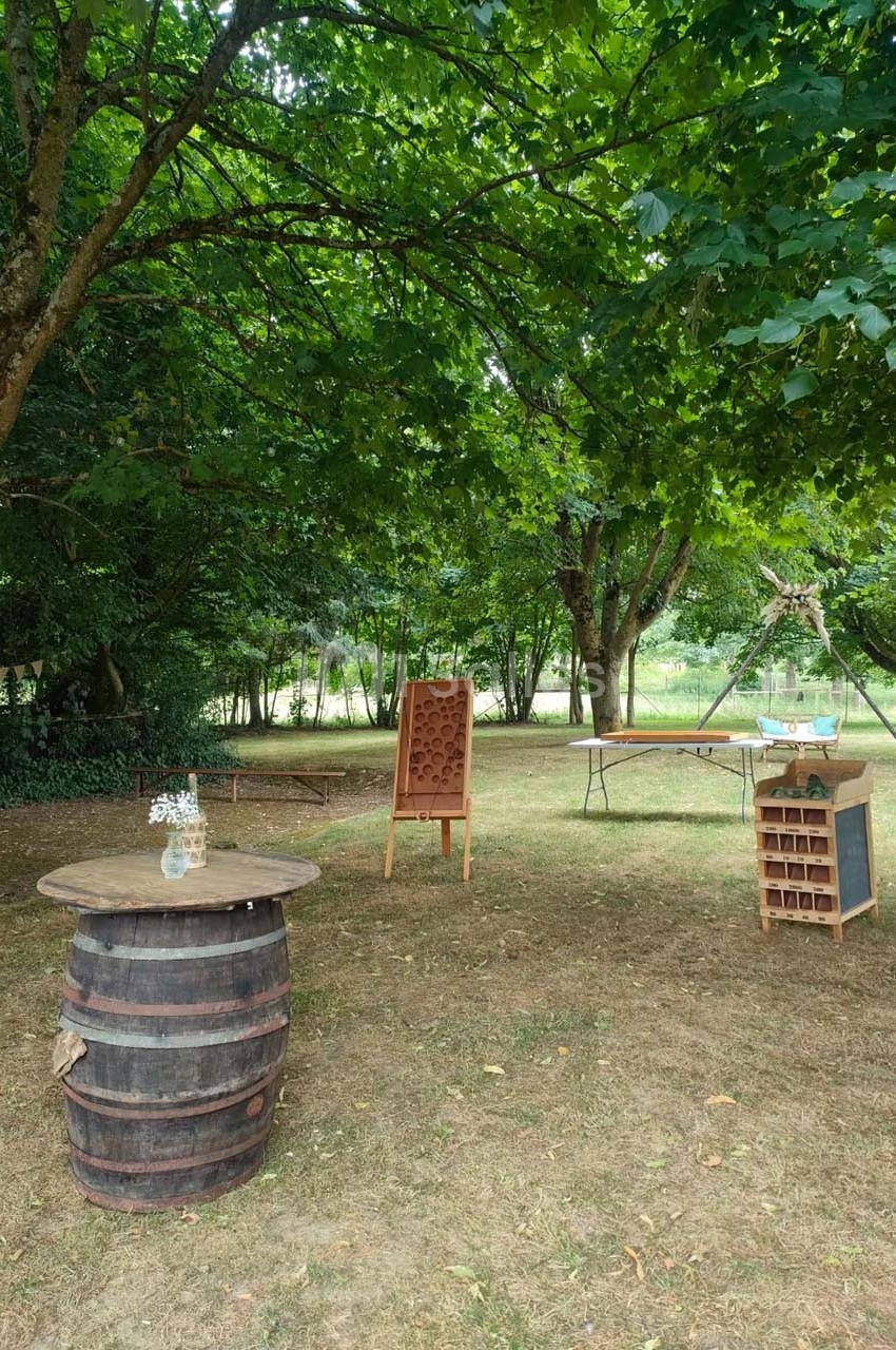 Espace extérieur avec tonneau en bois, présentoirs en bois et tables disposés sous des arbres dans un cadre naturel.
