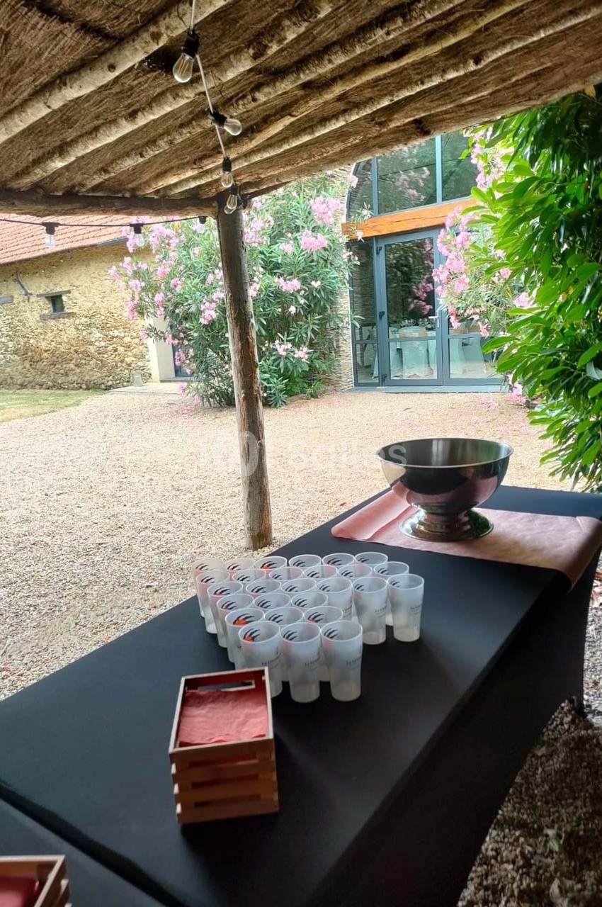 Table avec verres en plastique alignés et une grande coupe métallique, sous un abri en bois dans un jardin fleuri.