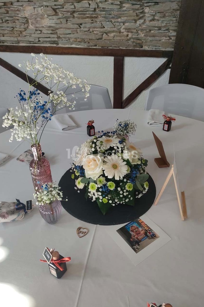 Centre de table avec bouquet de fleurs blanches et bleues, décorations et photo posés sur une nappe blanche.