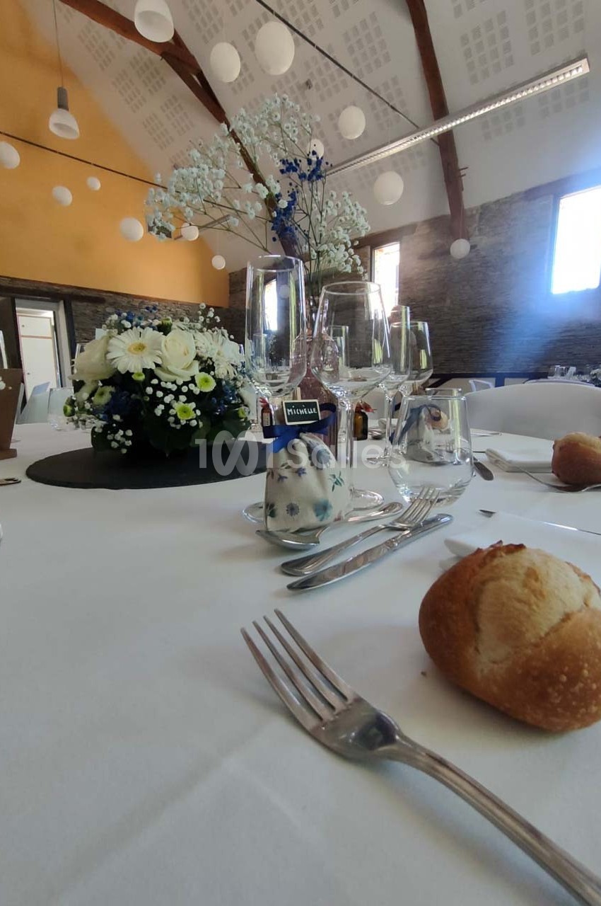 Table dressée avec nappes blanches, fleurs, verres, couverts et petits pains dans une salle lumineuse au plafond en poutres…
