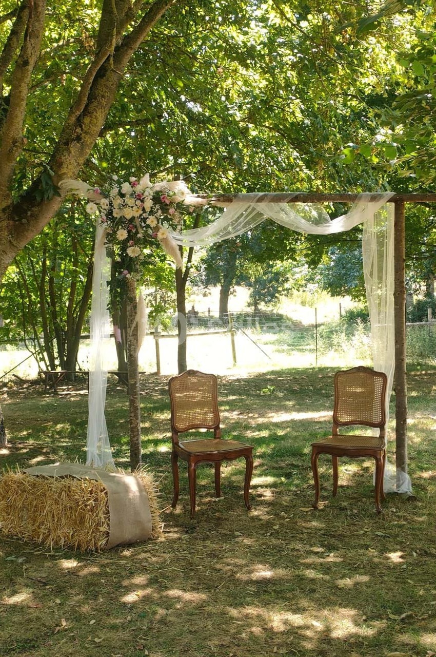 Arche en bois décorée de voilages et de fleurs, avec deux chaises en osier et une botte de paille, dans un jardin ombragé.