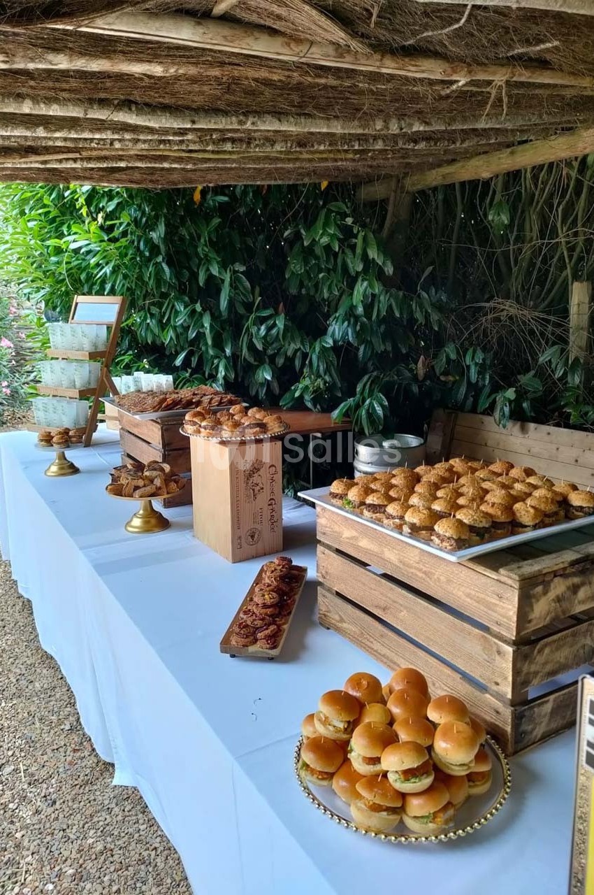 Buffet en extérieur avec mini-burgers, viennoiseries et desserts disposés sur une table blanche sous une pergola.