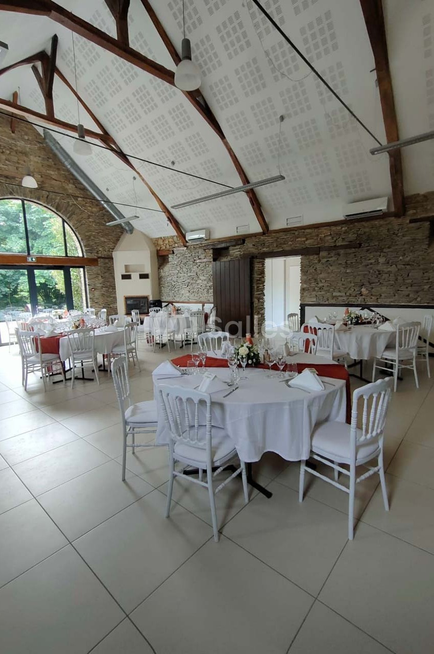Salle lumineuse avec tables rondes dressées, chaises blanches et murs en pierre, vue sur un jardin à travers de grandes…