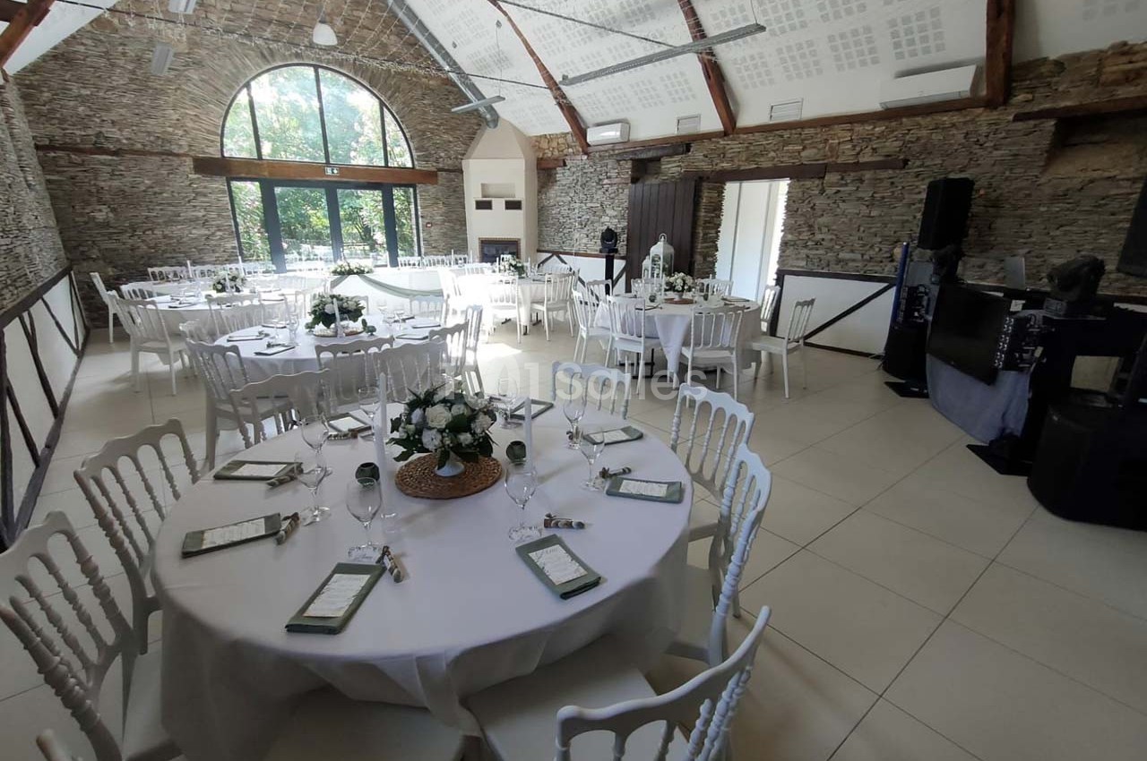 Salle de réception lumineuse avec tables rondes dressées, décorées de fleurs blanches et chaises blanches.