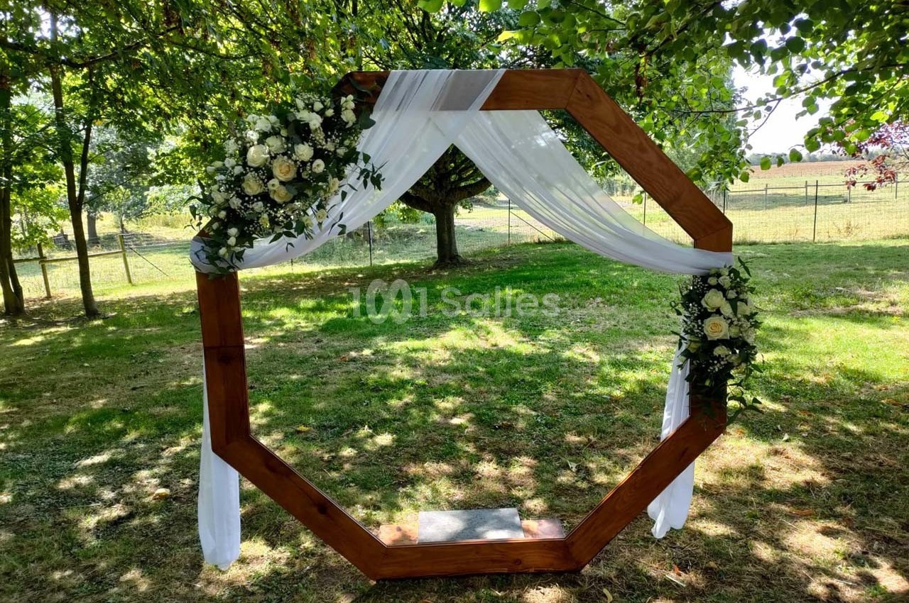 Arche octogonale en bois décorée de fleurs blanches et de voilages, installée dans un jardin verdoyant.