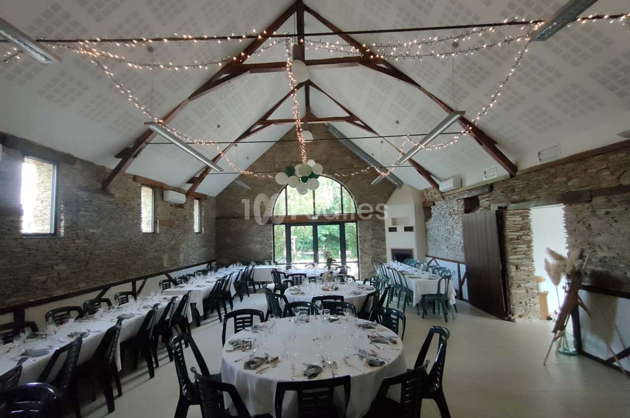 Salle de réception avec murs en pierre, tables rondes dressées et guirlandes lumineuses suspendues au plafond.