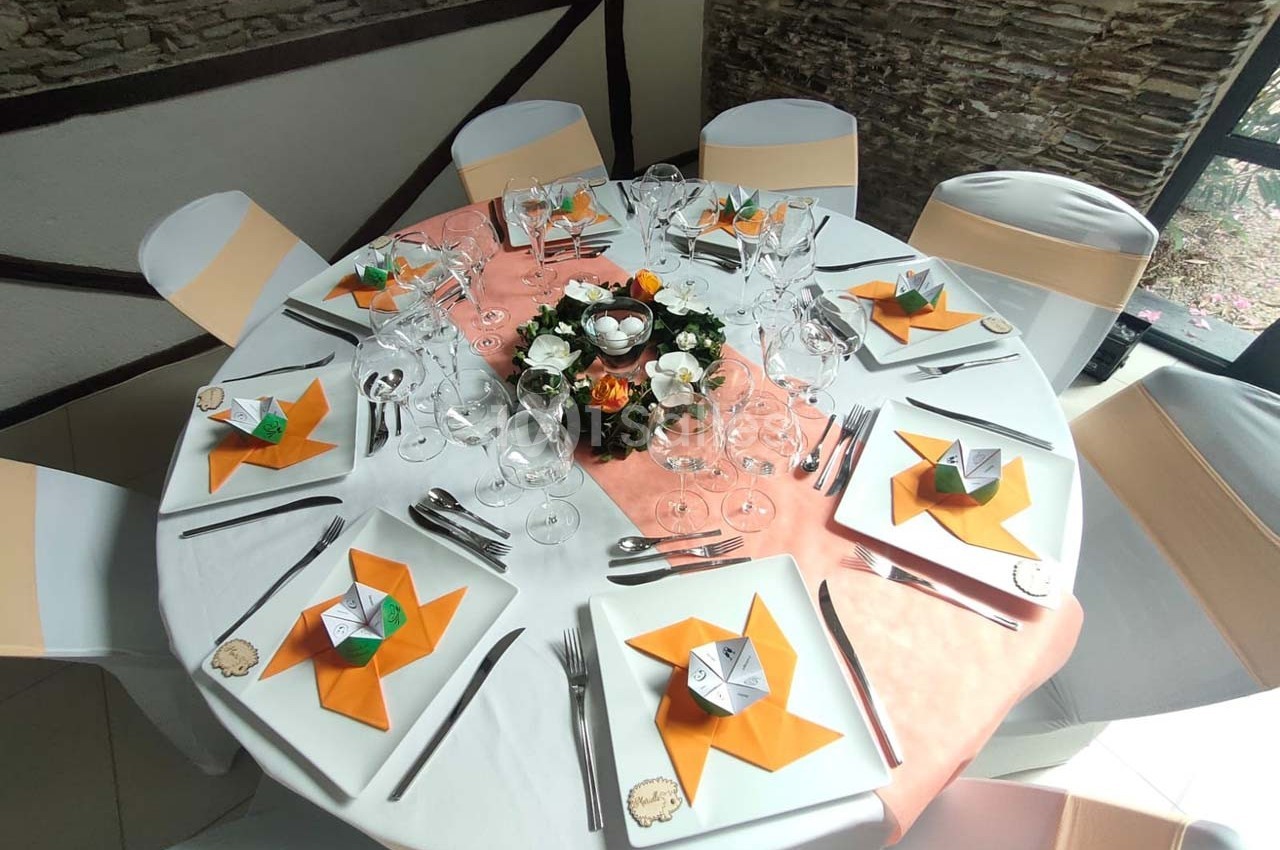 Table ronde dressée pour un repas, avec nappes blanches, serviettes pliées en étoile orange et décoration florale centrale.