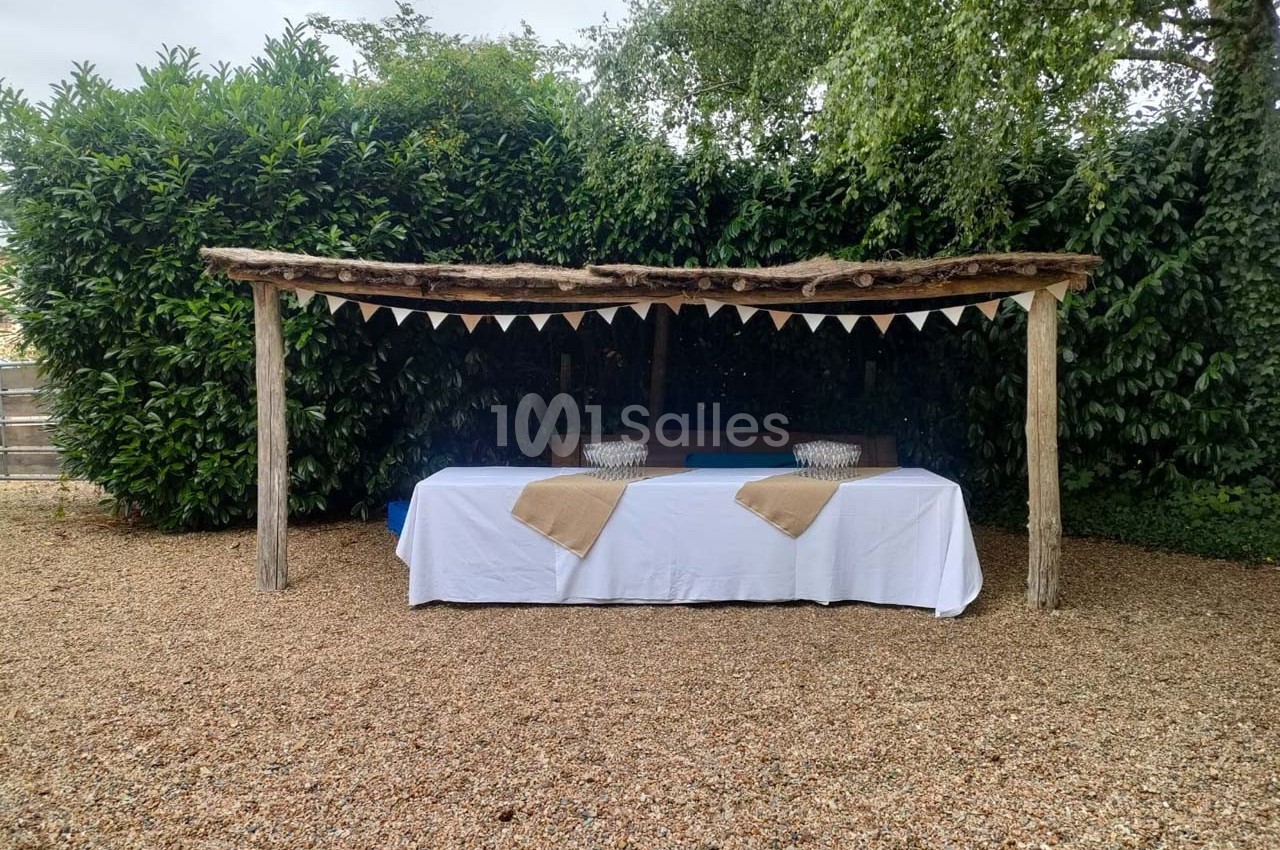 Table recouverte d'une nappe blanche sous une pergola en bois, décorée de fanions, sur un sol en gravier.