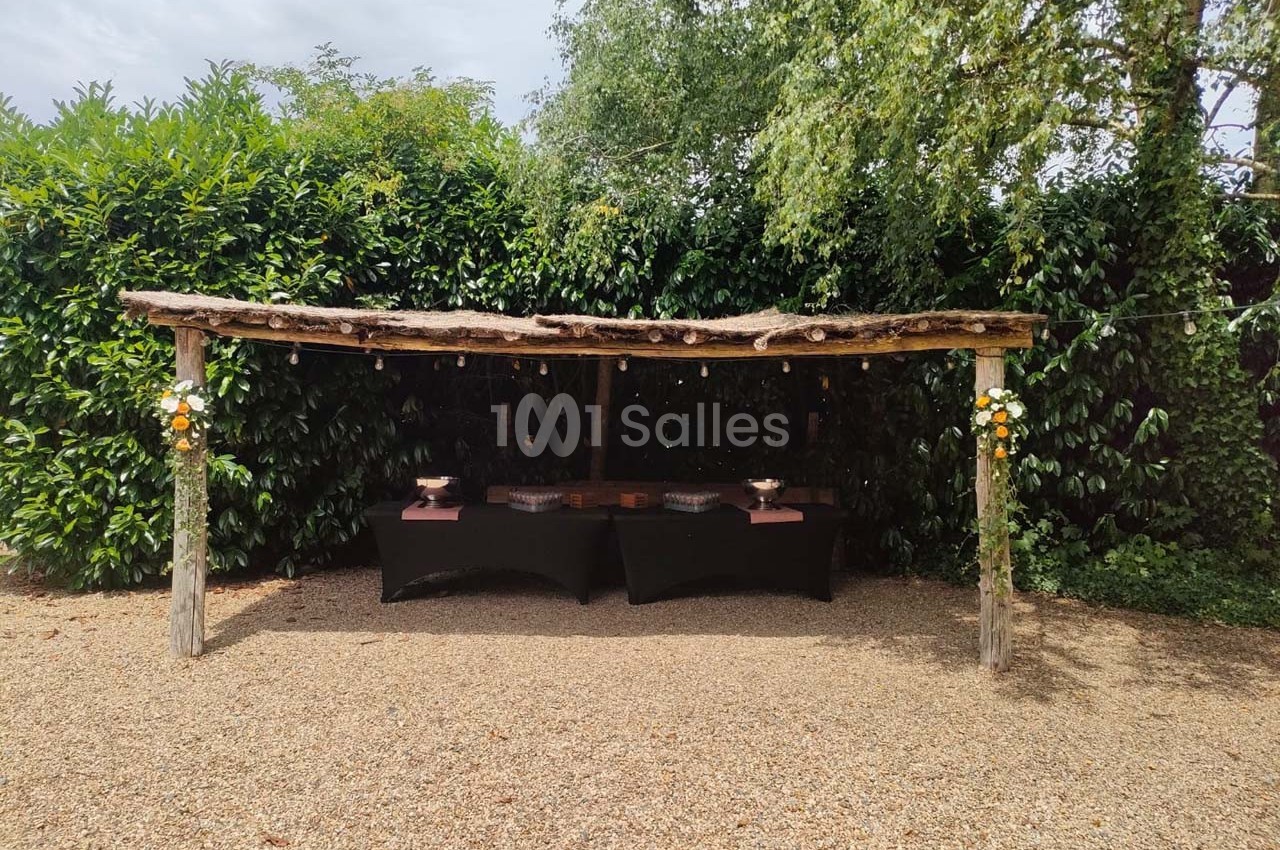 Abri en bois rustique avec tables noires dressées, entouré de végétation et décoré de fleurs.