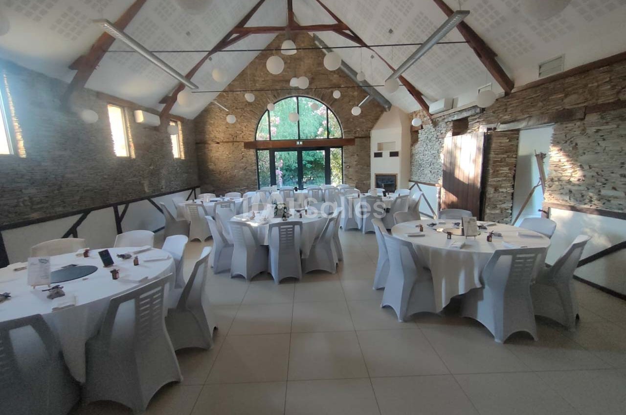 Salle de réception lumineuse avec tables rondes dressées et chaises blanches, murs en pierre et grande baie vitrée.