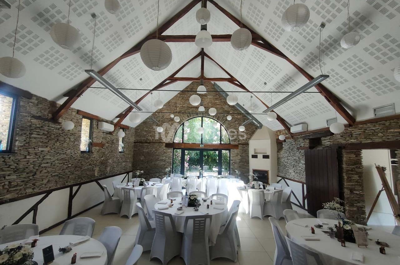 Salle de réception lumineuse avec murs en pierre, tables rondes dressées et lanternes suspendues au plafond.