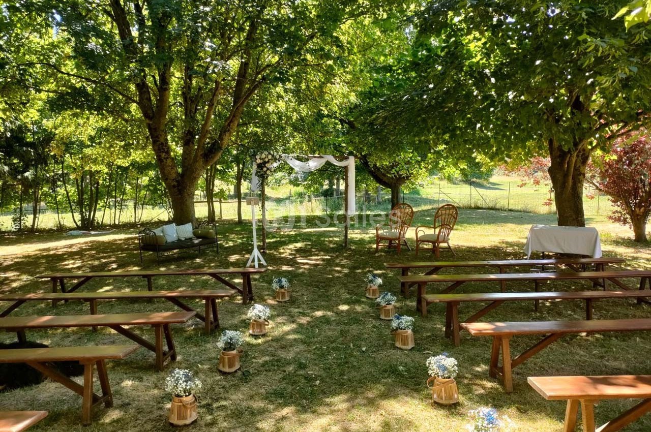 Cérémonie en plein air avec bancs en bois, arche décorée et chaises sous des arbres dans un cadre verdoyant.