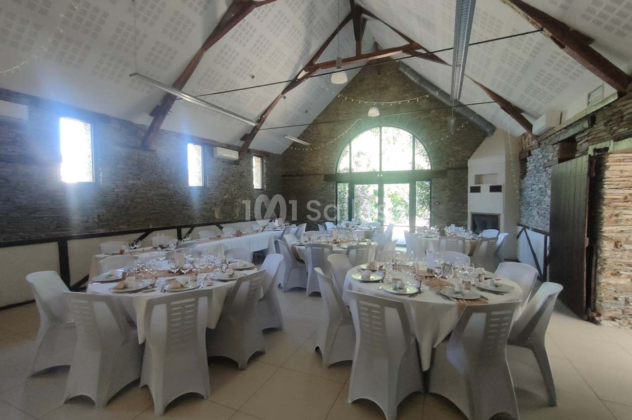 Salle de réception lumineuse avec des tables rondes dressées, chaises blanches et murs en pierre apparente.