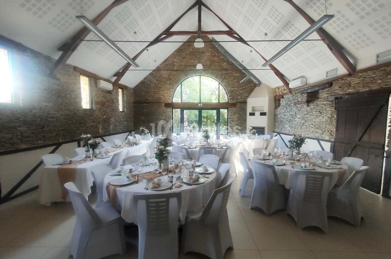 Salle de réception lumineuse avec tables rondes dressées, nappes blanches et décorations florales, murs en pierre apparente.