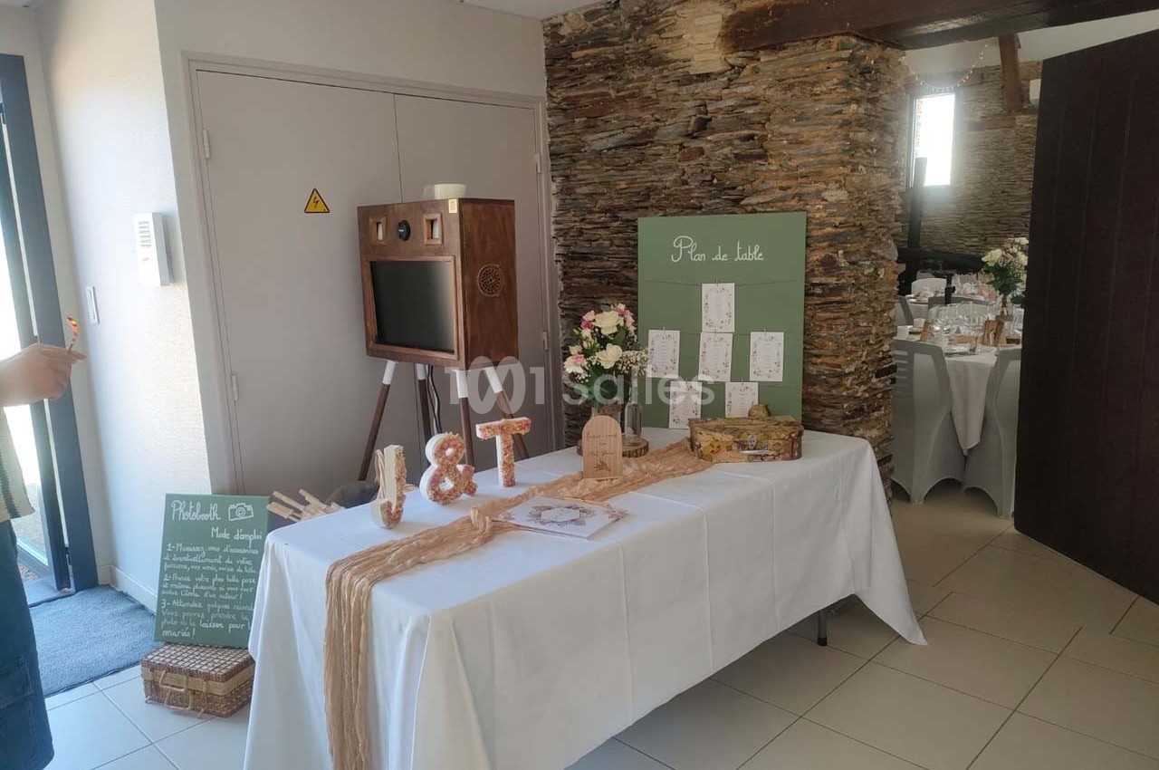 Table décorée pour un mariage avec urne, plan de table, fleurs et photobooth en bois dans une salle lumineuse.