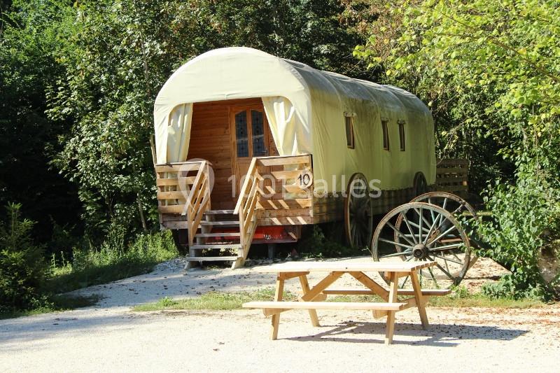 Roulotte en bois avec toit arrondi, entourée de végétation, avec une table de pique-nique et une roue décorative.