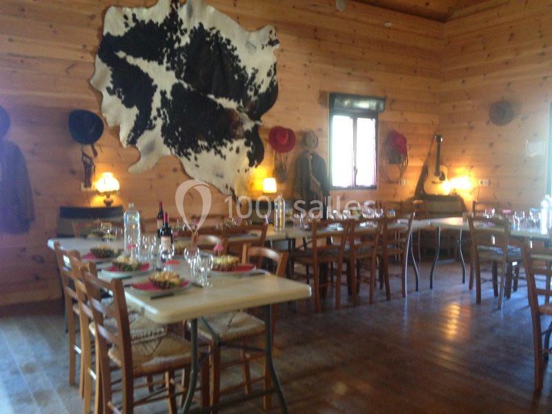 Salle en bois rustique avec tables dressées pour un repas, décorée de chapeaux, d'une peau de vache et d'une guitare.