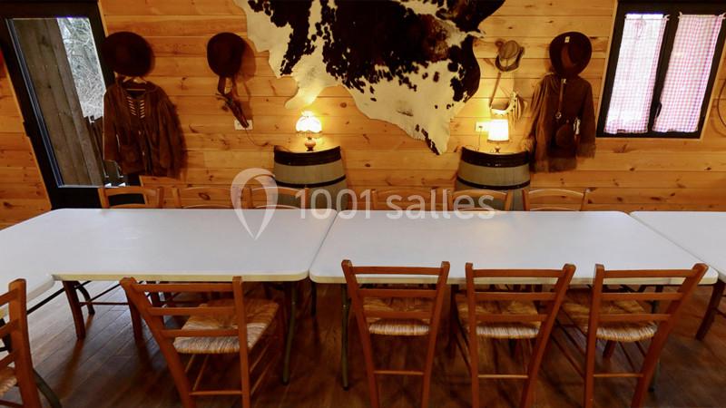 Salle rustique avec murs en bois, tables blanches, chaises en bois, décorations western et peau de vache accrochée au mur.