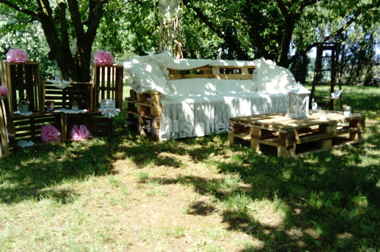 Mobilier en palettes décoré de tissu blanc et de fleurs roses, disposé dans un jardin ombragé.