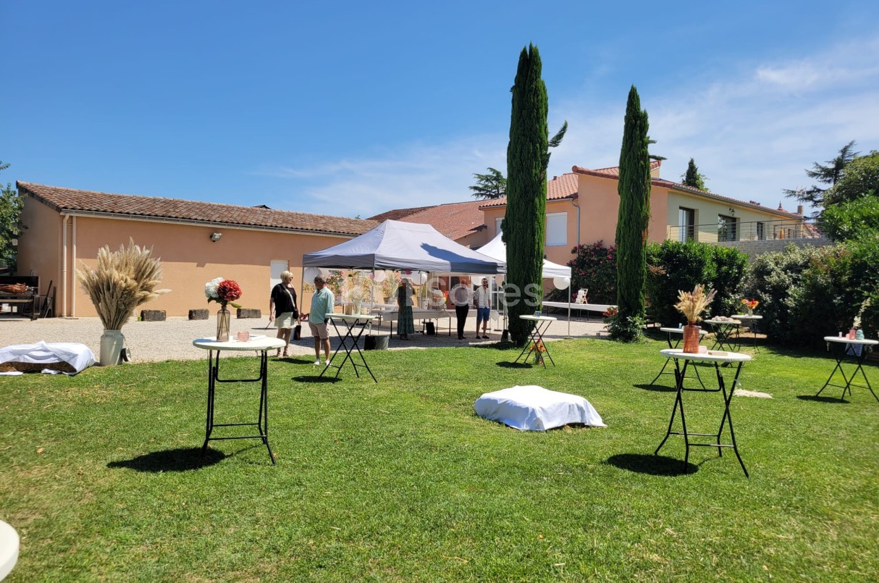 Jardin avec tables hautes décorées, tentes blanches et invités rassemblés près d'une maison sous un ciel ensoleillé.
