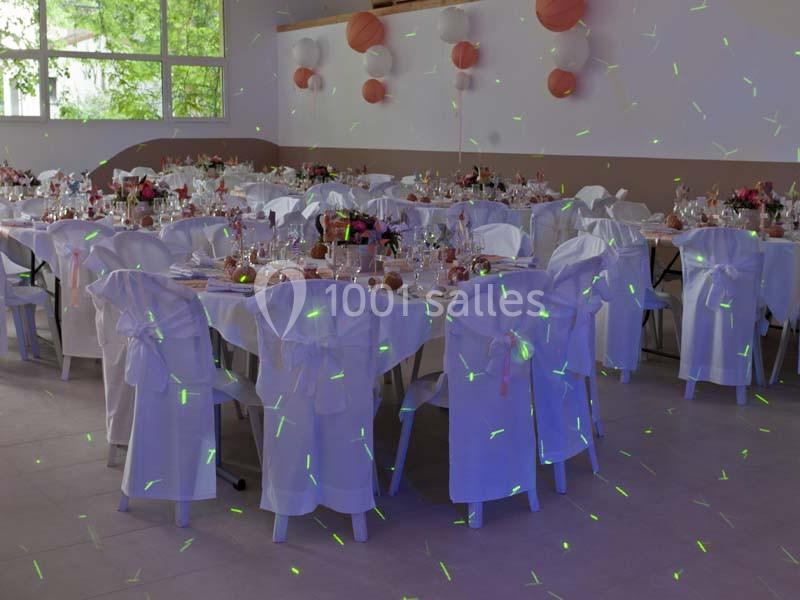 Salle de réception décorée avec des tables rondes, nappes blanches, chaises ornées de rubans et lumières colorées.