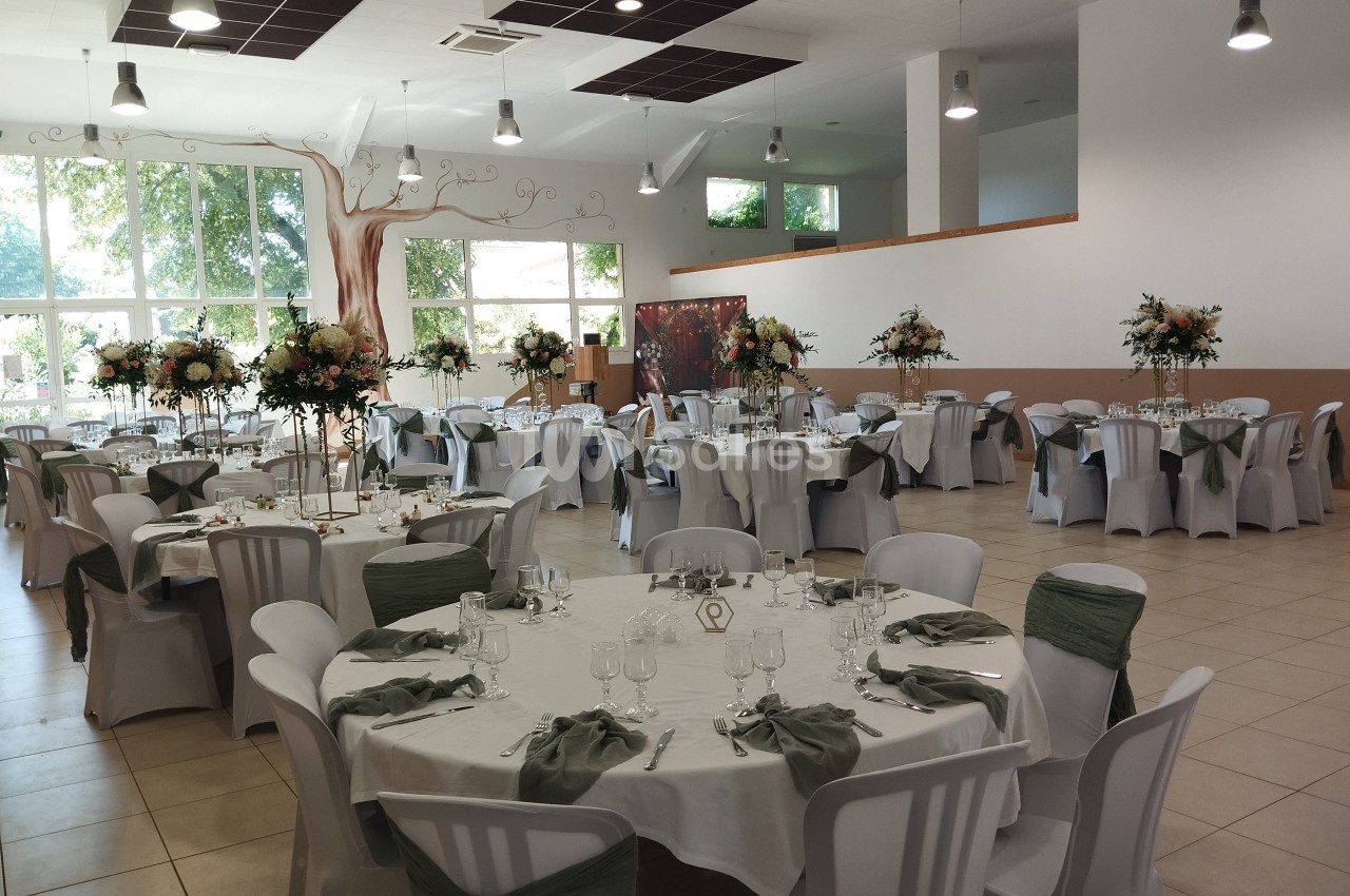 Salle de réception décorée avec des tables rondes, nappes blanches, chaises ornées de nœuds verts et centres de table…