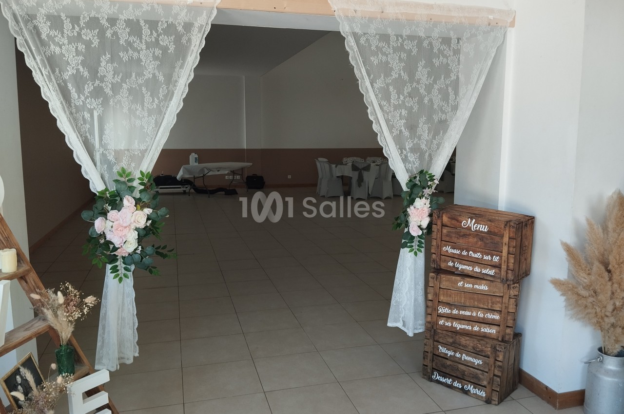 Salle de réception décorée avec rideaux en dentelle, fleurs, menu sur caisses en bois et chaises blanches au fond.