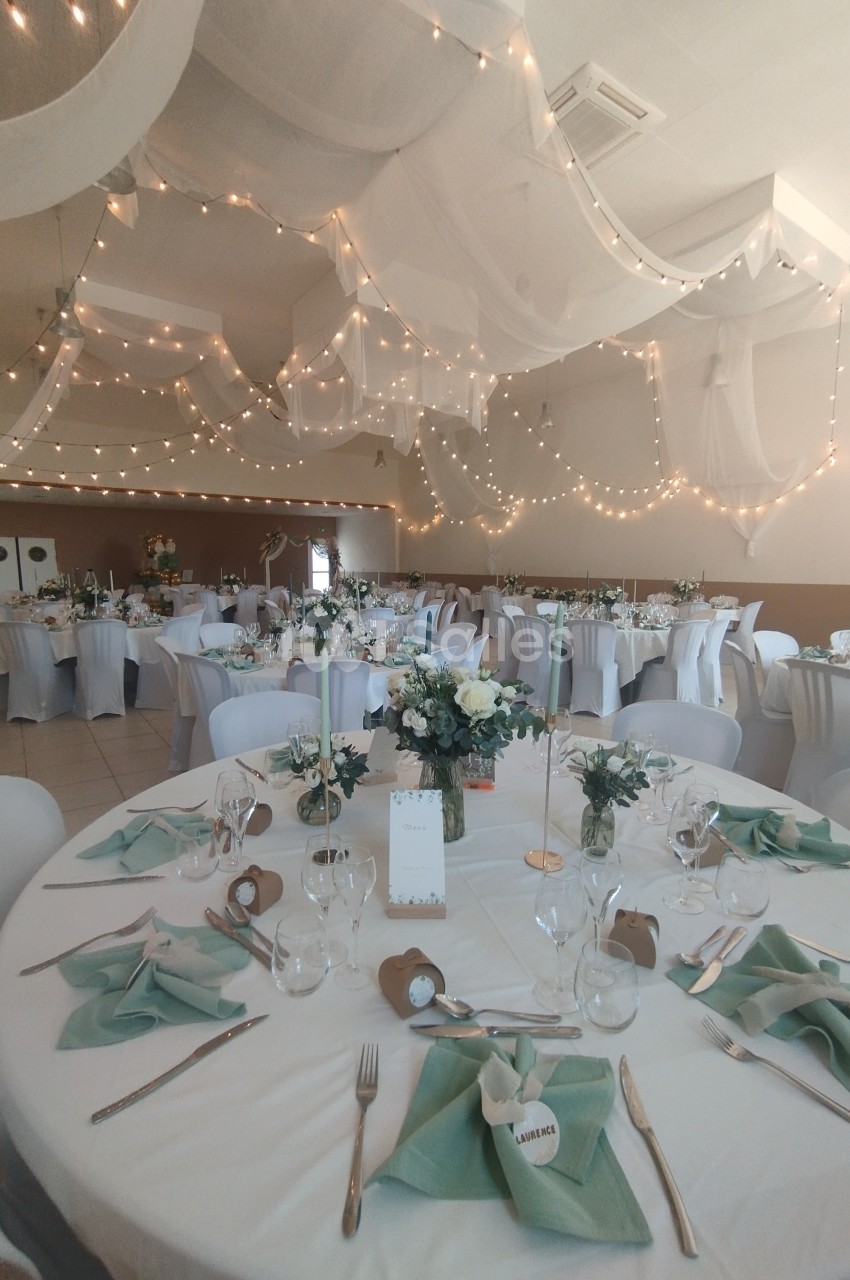 Salle de réception décorée avec des tables rondes, nappes blanches, serviettes vertes et guirlandes lumineuses suspendues.