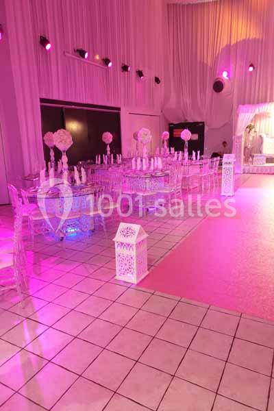 Salle de réception décorée avec des tables transparentes, des fleurs roses et un éclairage rose tamisé.