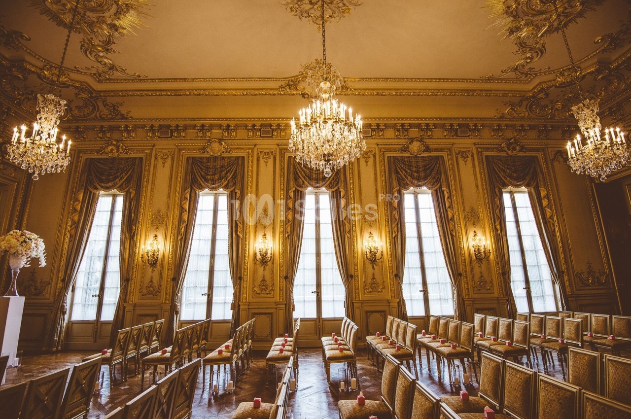 Salle de réception élégante avec lustres, hauts plafonds ornés et rangées de chaises alignées devant de grandes fenêtres.