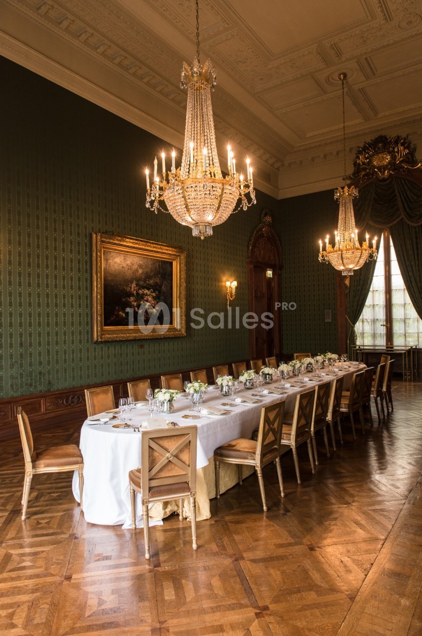 Grande salle élégante avec une longue table dressée, entourée de chaises en bois, éclairée par des lustres en cristal.
