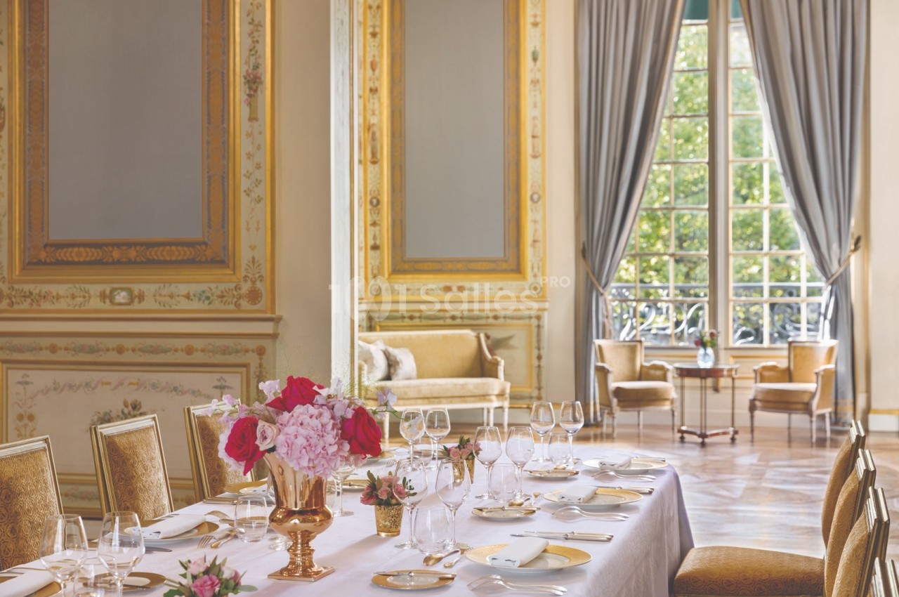 Salle élégante avec table dressée, décorée de fleurs, moulures dorées, grandes fenêtres et rideaux ouverts sur un jardin.