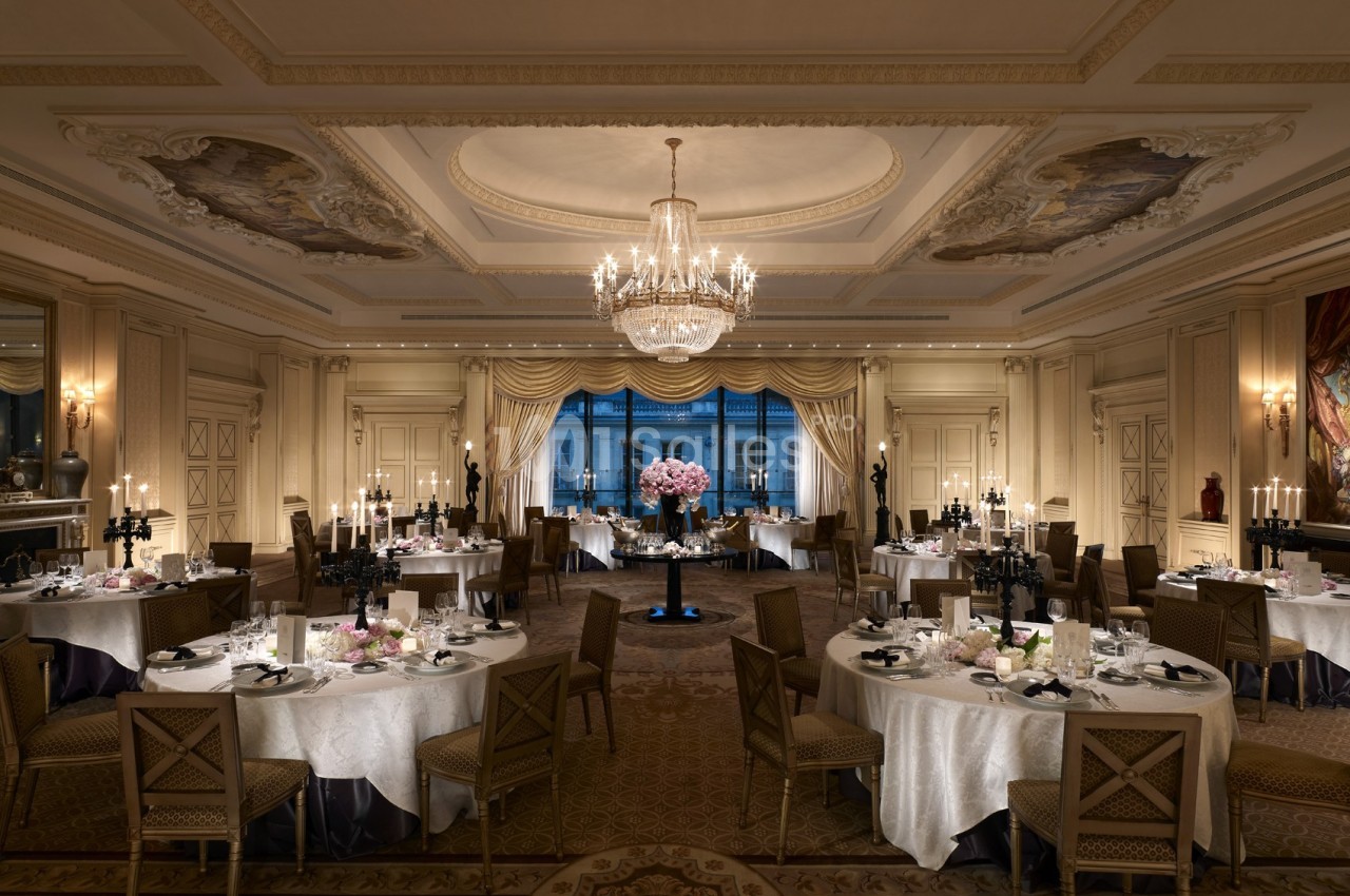 Salle de réception élégante avec tables rondes dressées, lustre central et grandes fenêtres donnant sur l'extérieur.